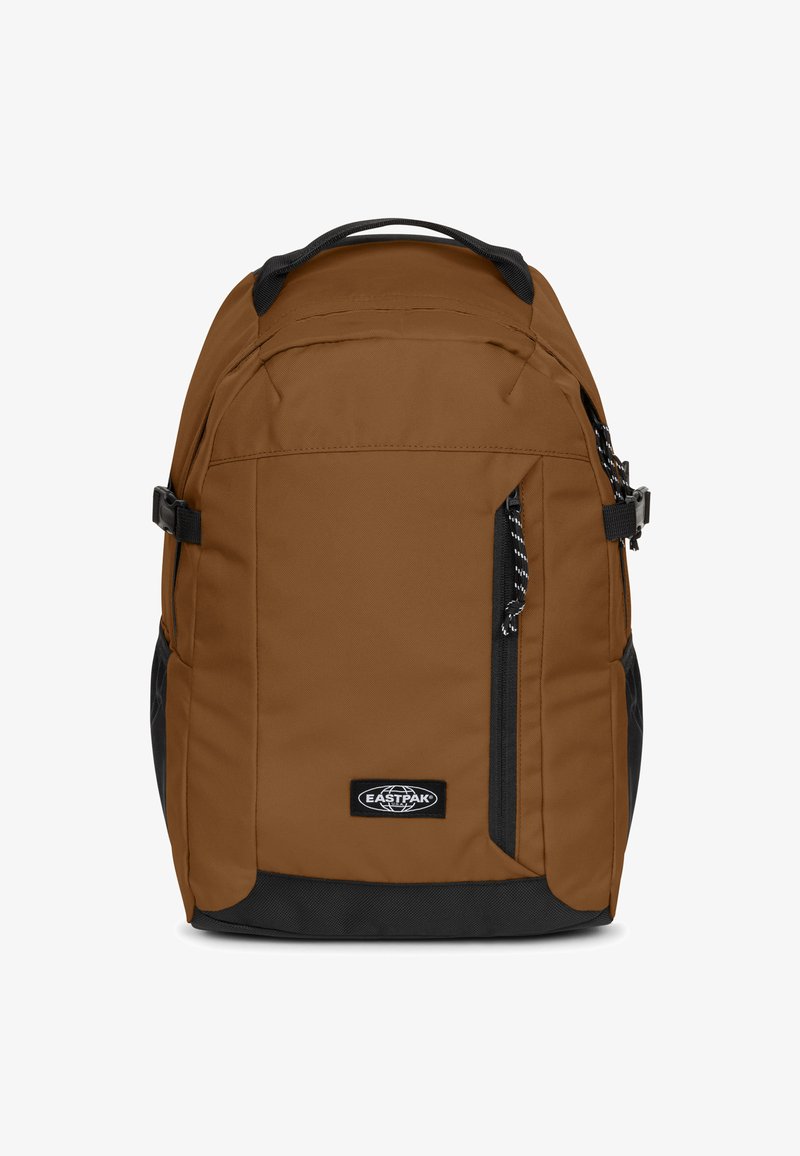 Mochila de tela marrón con detalles negros, cremalleras y un bolsillo delantero. Cuenta con correas ajustables y un asa superior para llevar.