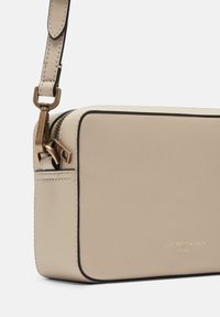 Beige Leder Crossbody-Tasche mit strukturierter Oberfläche, goldenen Beschlägen und Reißverschlussverschluss. Verfügt über ein dezentes Logo und kontrastierenden schwarzen Besatz.