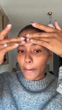 Grå, strikket genser med ribbet hals. Hender påfører hudpleie i pannen. Naturlig lys belyser ansiktet.