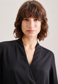 Schwarze Wickelbluse mit einem V-Ausschnitt, Knopfdetails an der Brust und glatter Textur. Kurze Ärmel und ein elegantes, minimalistisches Design.