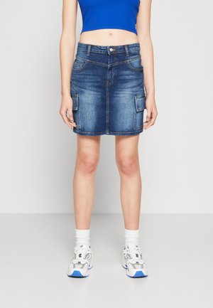 Donna con un top corto blu, una gonna cargo in denim con tasche laterali, calze bianche e sneakers bianche su uno sfondo semplice.