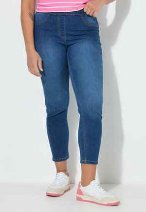 Person, die geschnittene blaue Jeans, weiße und pinke Sneakers sowie ein pink gestreiftes Oberteil trägt, vor einem weißen Hintergrund steht.