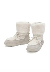 Botas de tobillo con suela gruesa en un suave color crema, con una parte superior de felpa, cordones en la parte delantera y un puño doblado. La superficie texturizada aporta interés visual.