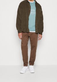 Braunes Cordhemd über einem türkisfarbenen Sweatshirt mit weißem Aufdruck, kombiniert mit braunen Hosen und weißen Sneakers, vor einem weißen Hintergrund stehend.