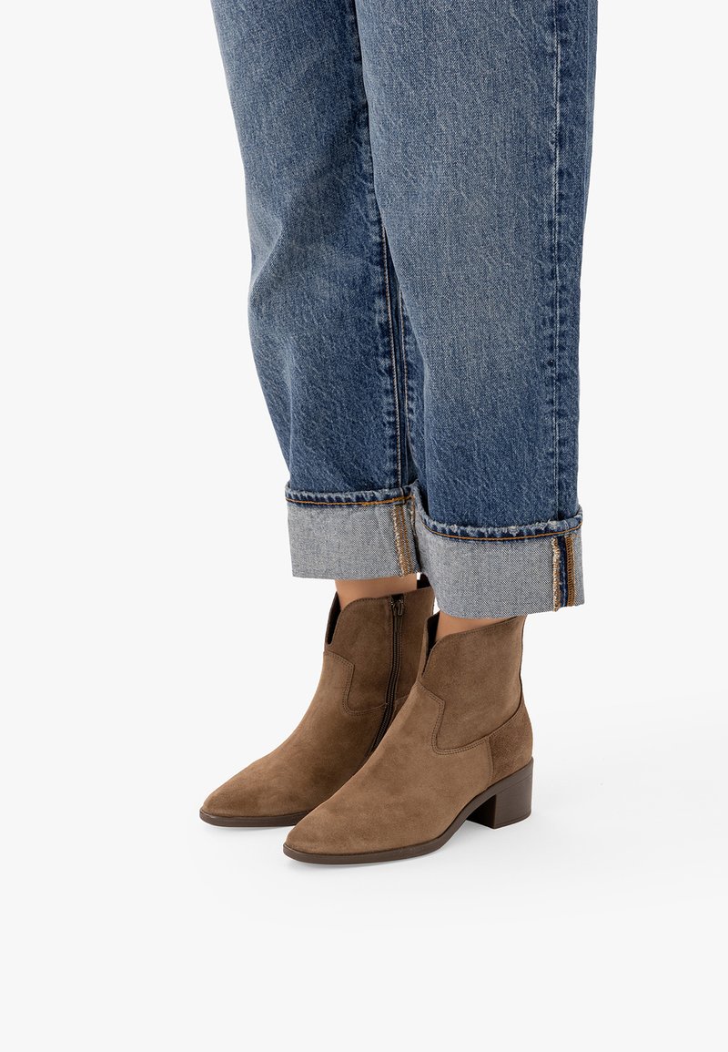 Bottines en daim marron avec fermetures éclair latérales, portées avec un jean bleu retroussé sur fond blanc.