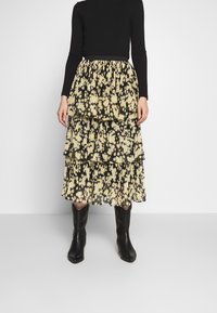 Una persona lleva una blusa negra de manga larga y una falda de volantes con patrones florales amarillos. Botas negras completan el atuendo sobre un fondo sencillo.