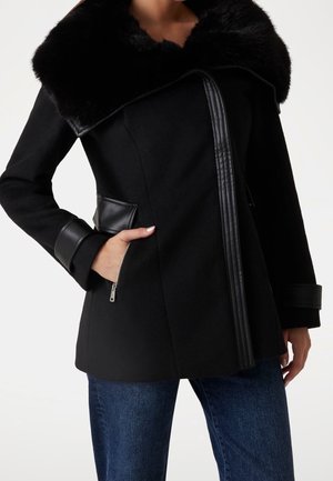 Femme portant un manteau noir avec un grand col en fourrure, des détails en cuir, des poches zippées et un jean bleu.