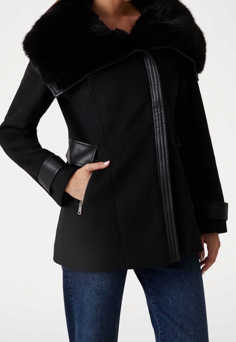 Femme portant un manteau noir avec un grand col en fourrure, des détails en cuir, des poches zippées et un jean bleu.