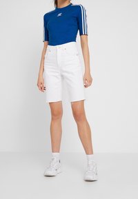 Blå kortärmad topp med Adidas-logotyp, tillsammans med vita knälånga denimshorts med fransade kanter och vita sneakers.