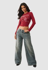 Crop top rouge à manches longues avec imprimé graphique, associé à un jean bleu déchiré à jambes larges. Des talons noirs complètent la tenue.