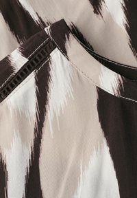 Nærbillede af stof med abstrakt sort, hvid og beige mønster og detaljeret søm langs en kant.