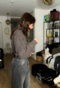 Γυναίκα με μακριά σκούρα μαλλιά, ντυμένη με γκρι πουλόβερ και τζιν, στέκεται σε ένα φορτωμένο σαλόνι και κοιτάζει το τηλέφωνό της.