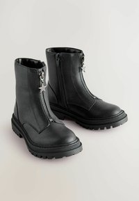 Bottines noires avec semelles épaisses, zips avant avec des tirettes en forme d'étoile et zips latéraux sur un fond clair.