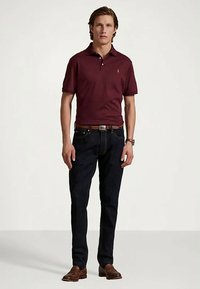 Un homme se tenant debout, portant un polo bordeaux, un jean bleu foncé, une ceinture en cuir marron, des chaussures marron et une montre au poignet, devant un fond clair.