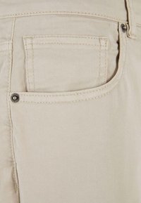 Pantalones de algodón beige con un bolsillo trasero con un remache metálico, textura sutil y detalles de costura visibles.