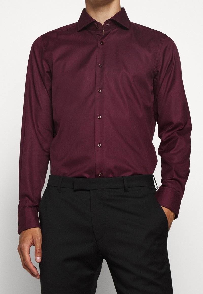 Homme portant une chemise ajustée bordeaux à manches longues rentrée dans un pantalon noir, une main dans la poche, sur fond uni.