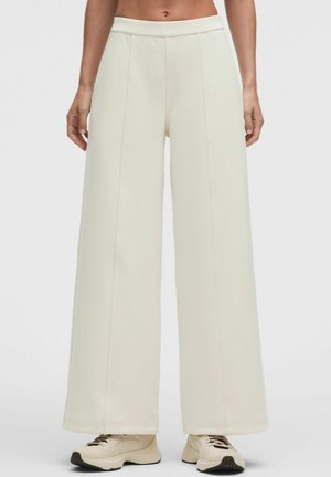 Donna che indossa pantaloni bianchi a vita alta e gamba larga e sneakers beige, in piedi davanti a uno sfondo bianco uniforme.