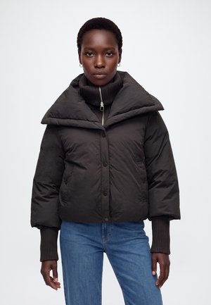 Femme portant une veste matelassée noire avec un large col par-dessus un pull zippé côtelé noir et un jean bleu, debout devant un fond uni.