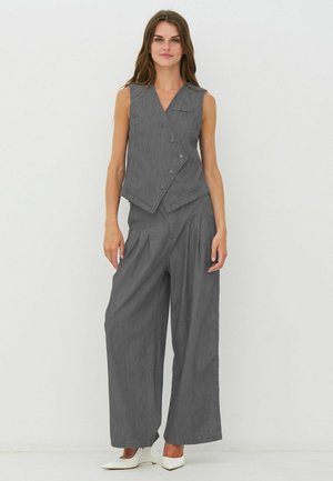 Femme portant un gilet gris sans manches boutonné et un pantalon large assorti avec des talons pointus blancs, debout devant un fond blanc.