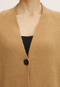 Cárdigan de punto en lana color beige con un profundo escote en V y un botón negro. Presenta textura acanalada y un ligero diseño de hombros caídos.