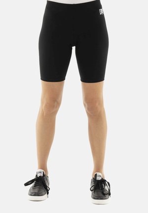 Personne portant un short noir moulant jusqu'aux genoux et des baskets noires avec semelles blanches et étiquettes "LEONE" sur les languettes.