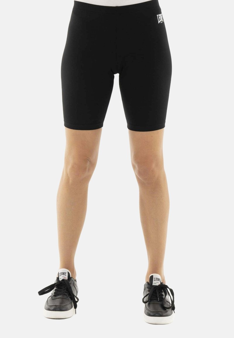 Personne portant un short noir moulant jusqu'aux genoux et des baskets noires avec semelles blanches et étiquettes "LEONE" sur les languettes.