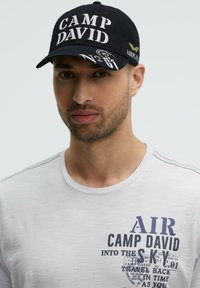 Navy cap with "CAMP DAVID" embroidered in white, curved brim, and yellow accent. Worn with a light-colored T-shirt featuring printed text.