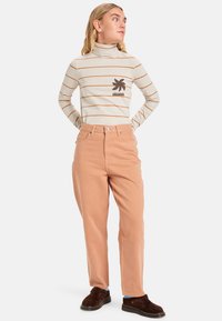 Un pull à col roulé crème à rayures avec un graphique marron foncé, associé à un pantalon rose pâle fuselé et des chaussures brunes à enfiler.