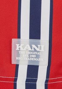 Raudona medžiaga su tamsiai mėlynomis ir baltomis vertikaliomis juostelėmis, kuriame yra aiškus KANI logotipo ženkliukas su tekstu "ORIGINALAS EST. 1989 REG. PREKĖS ŽENKLAS."