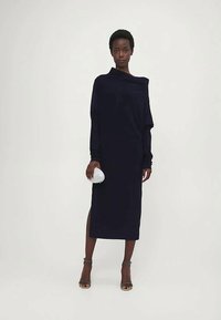 Nő egy hosszú ujjú, vállára lógó fekete midi ruhában oldalsó hasítékkal, kis fehér táskával a kezében és fekete nyitott orrú magassarkú cipőben.