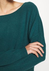 Türkis gestrickter Pullover mit einem weiten Bootsausschnitt, langen Ärmeln und einer weichen Textur, mit sichtbaren Stichen entlang der Kanten und Bündchen.