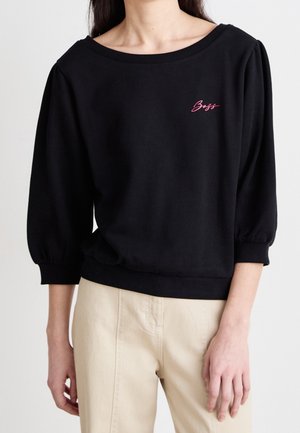 Vrouw draagt een zwart sweatshirt met driekwart pofmouwen en roze "Boss" tekst op de linkerborst, gecombineerd met beige broek.
