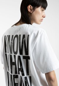 White cotton t-shirt with short sleeves, featuring large black distressed text on the back reading "KNOW WHAT I MEAN." Rounded neckline.