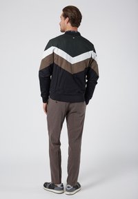 Giacca da uomo con motivo a zig zag nei colori nero, bianco, verde e marrone. Tessuto testurizzato, polsini a costine. Pantaloni a quadri marroni e scarpe casual.