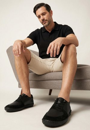 Mand i sort polotrøje og beige shorts, der sidder på en grå sofa, iført sorte afslappede sko med snørebånd, ser eftertænksomt nedad.