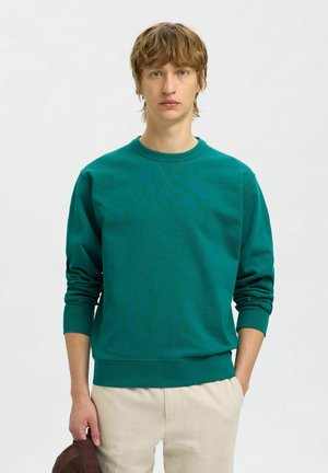 Teal sweatshirt met lange mouwen, ronde hals en geribbelde manchetten. Gecombineerd met lichtgekleurde broek, vastgehouden met een bruin voorwerp in de hand.