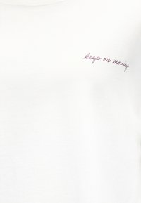 Λευκό ύφασμα με μικρό, καλλιγραφικό μοβ κείμενο κοντά στην επάνω δεξιά γωνία που γράφει «συνέχισε να κινείσαι».