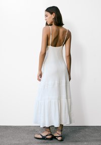 Robe longue blanche avec des bretelles fines ajustables, volants superposés au bas, tissu texturé et un dos légèrement ouvert. Associée avec des sandales à lanières.