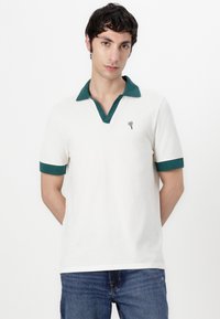 Biely polo tričko so zeleným golierom a manžetami; má malé logo srdca na hrudi; vyrobené z hladkej tkaniny.
