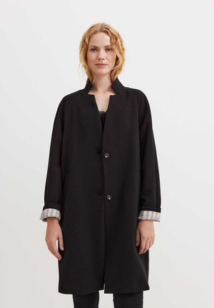 Cappotto nero con colletto alla mandarina, dotato di chiusura a pulsante, taglio over-size e dettagli delle maniche a strisce arrotolate. Tessuto dalla texture liscia.