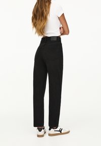 Femme aux longs cheveux portant un jean noir taille haute, un t-shirt court blanc et des baskets blanches avec des accents noirs, debout les bras croisés.