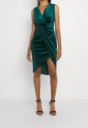 Femme portant une robe portefeuille sans manches en velours vert foncé avec un col en V et un ourlet asymétrique, associée à des escarpins noirs à bride cheville.