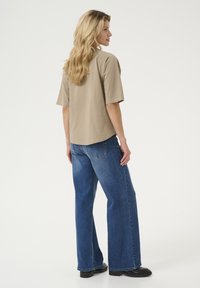 Camisa de manga corta beige con un corte relajado, combinada con jeans de pierna ancha azul oscuro, que tienen bolsillos traseros y un ligero desvanecimiento. Zapatos negros.