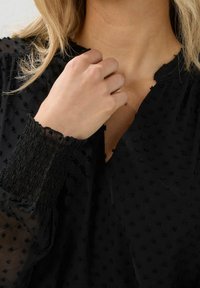 Primo piano di una persona che tocca il colletto indossando una blusa nera a maniche lunghe con texture, polsini arricciati e scollatura a V.