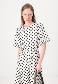 Vestido branco com bolinhas pretas; apresenta mangas curtas em balão e uma cintura com cinto. Tecido suave com um design de saia esvoaçante.