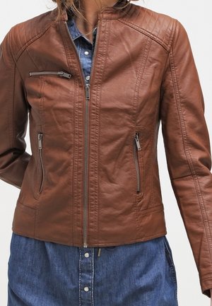 Frau trägt eine figurbetonte braune Lederjacke mit gesteppter Schulterpartie über einem blauen Denim-Kleid mit Knopfleiste.