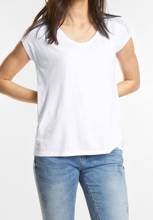 Biała koszulka z krótkim rękawem i okrągłym dekoltem, z teksturowanymi akcentami na ramionach, w połączeniu z jasnoniebieskimi dżinsami z delikatnymi przetarciami.