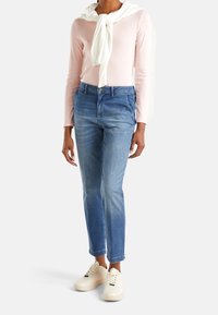 Femme portant une chemise rose pâle à manches longues avec un pull blanc noué autour des épaules, un jean bleu et des baskets blanches sur fond blanc.