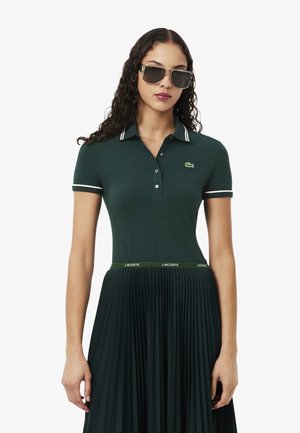 Donkergroene polo met witte accenten, voorzien van een klein logo op de borst, gecombineerd met een geplooide rok met een merkband in de taille.