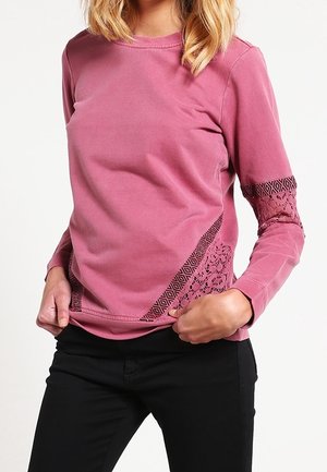 Vrouw die een lichtroze top met lange mouwen draagt met geometrisch kanten detail op de mouwen en zijkant, gecombineerd met een zwarte broek.